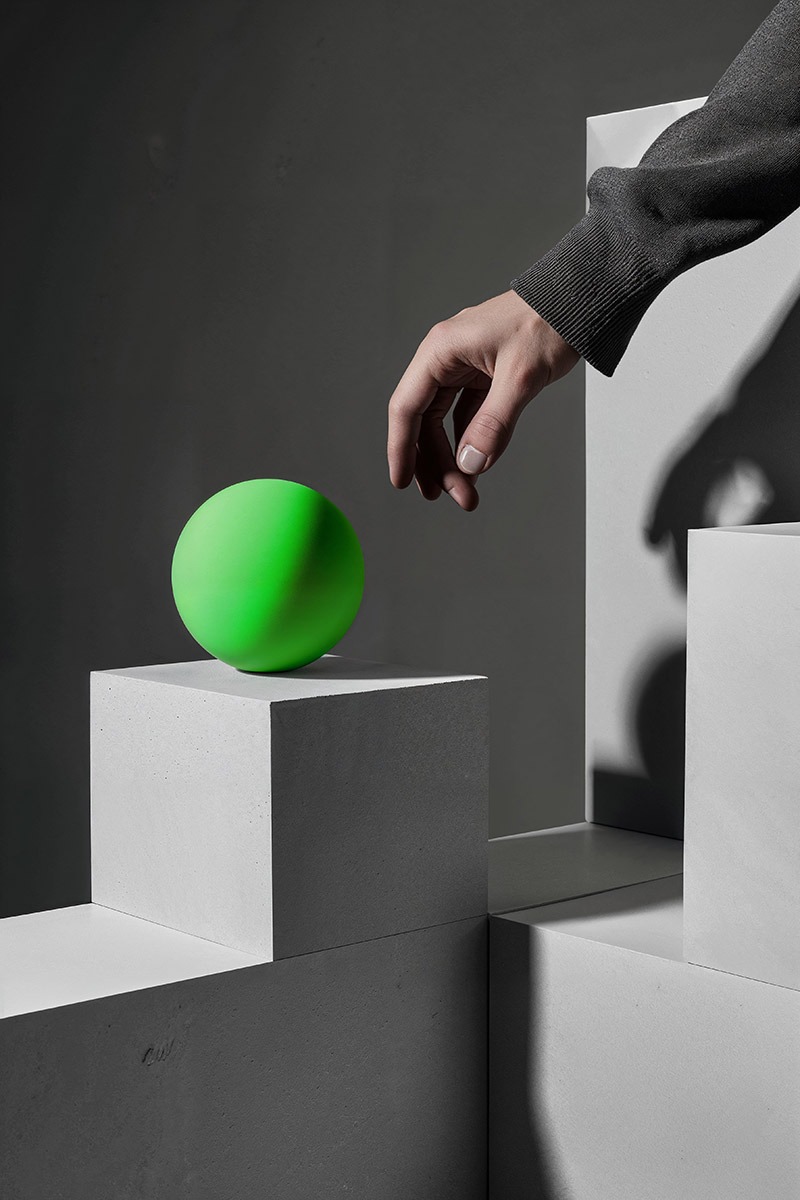 Nature morte en image de synthèse : une main effleure une sphère verte posée sur des blocs de béton